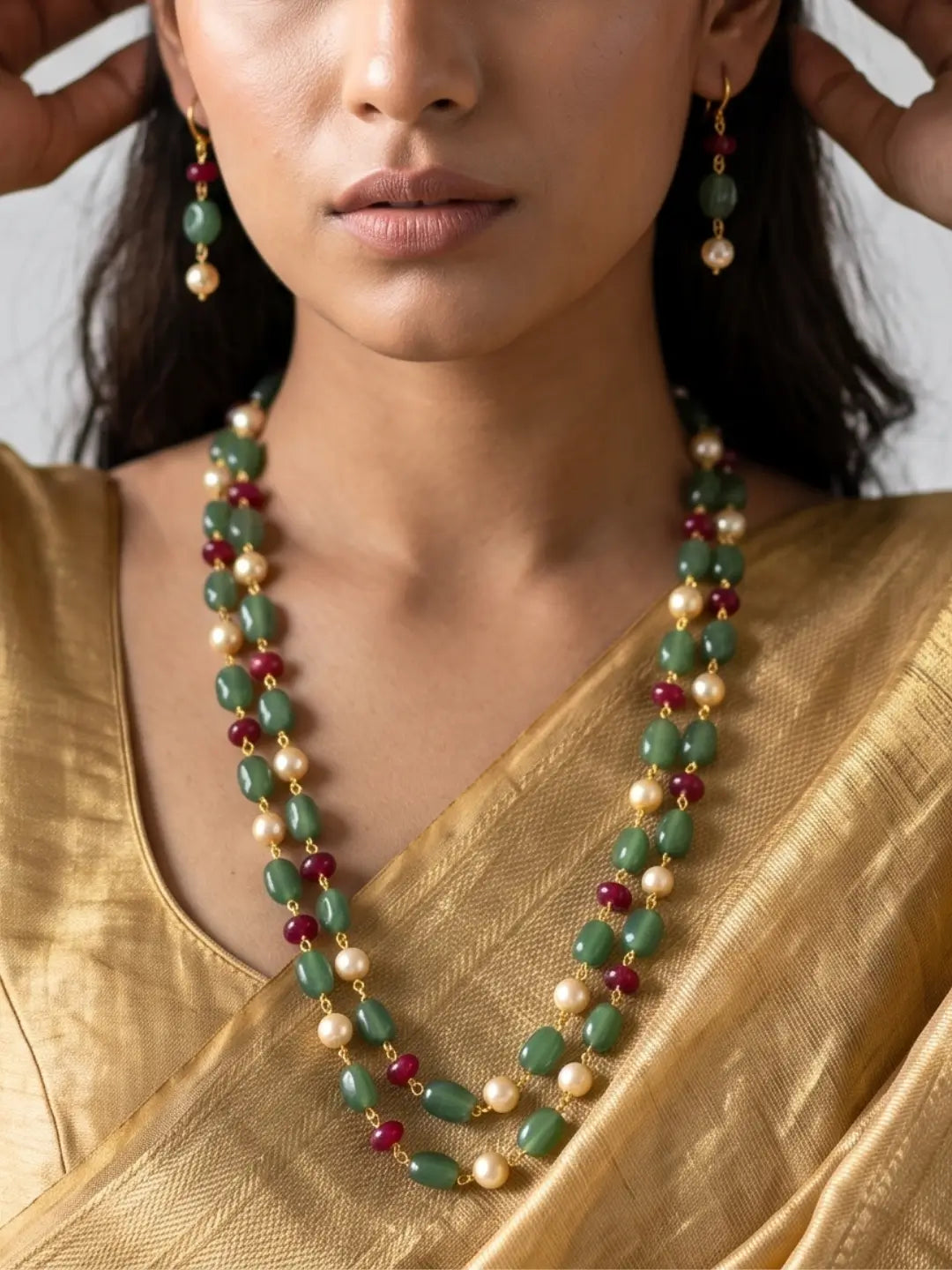 A close-up of a model wearing a double-strand green beaded necklace and matching drop earrings. The necklace features green stone beads, cream-colored pearls, and small red beads separated by gold-toned spacers, worn over a gold saree.