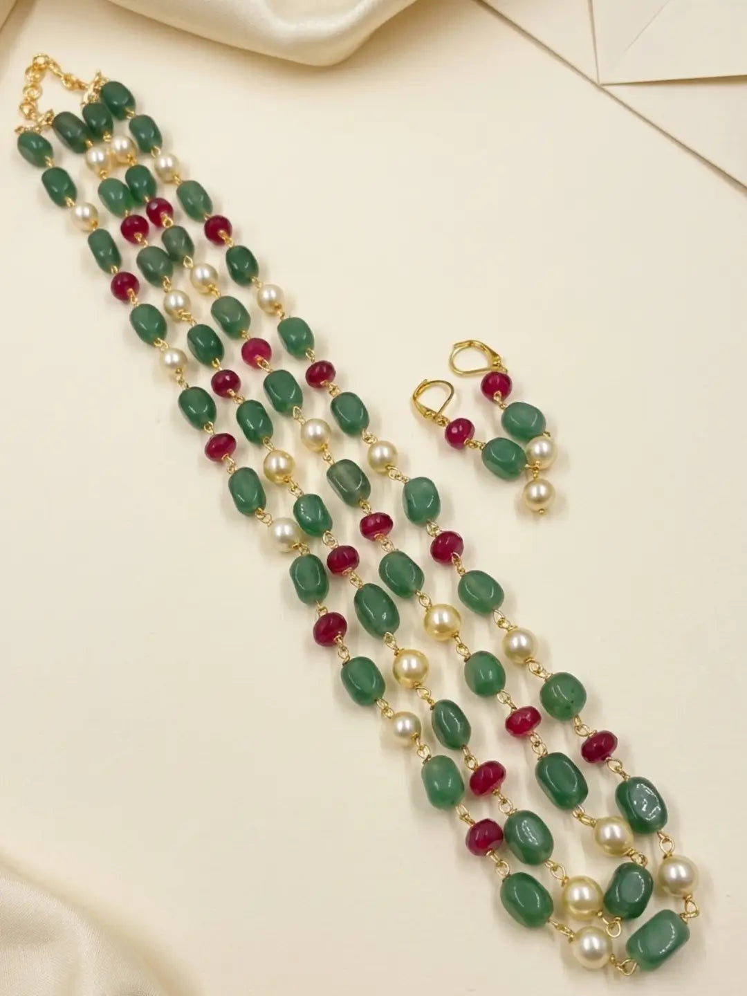 A gold-plated dual-strand necklace and matching drop earrings featuring green cabochon beads, white pearl beads, and small red faceted beads, arranged on a cream-colored fabric background.