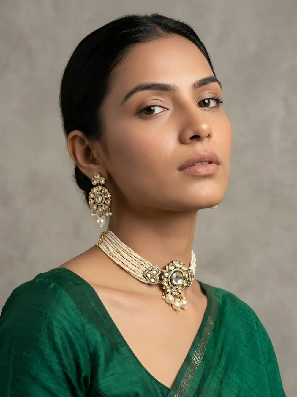 A woman wearing an ornate dual-toned polki choker necklace with pearl details and matching earrings, paired with a green blouse, against a neutral textured gray background.