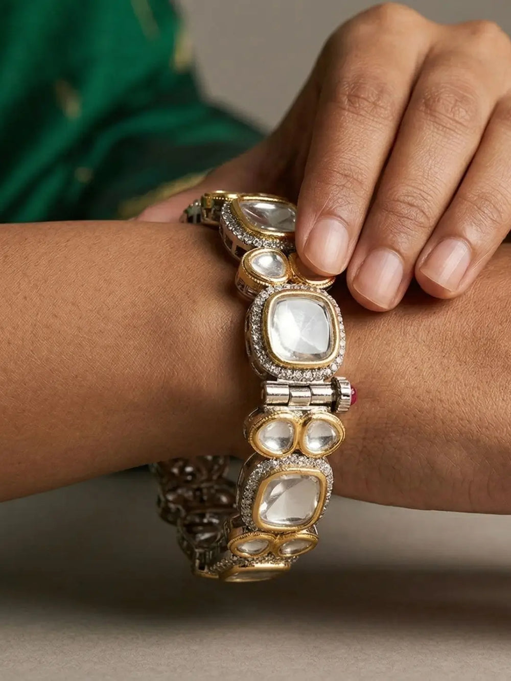 A close-up of a gold and silver-toned traditional Indian polki Kundan bracelet with clear stones, worn on a person's wrist with a hand resting on top.