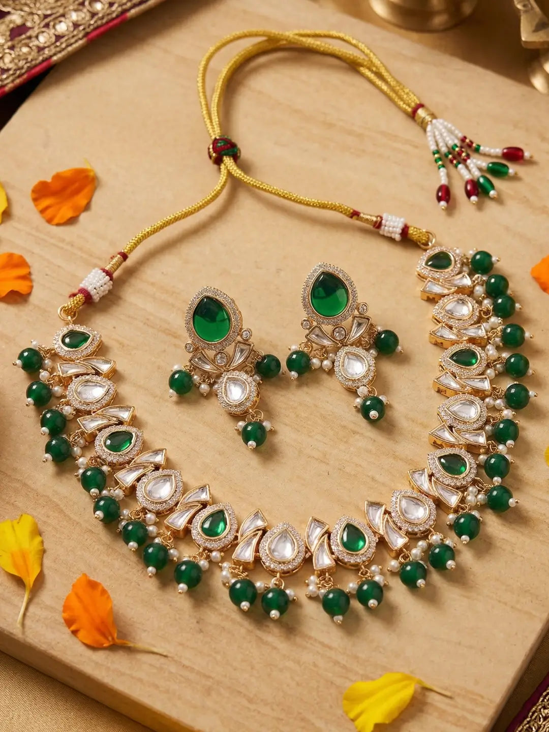 Gold-plated Indian Kundan jewelry set featuring a necklace and matching earrings with green stones and clear crystals, displayed on a wooden surface with decorative orange and yellow leaves.