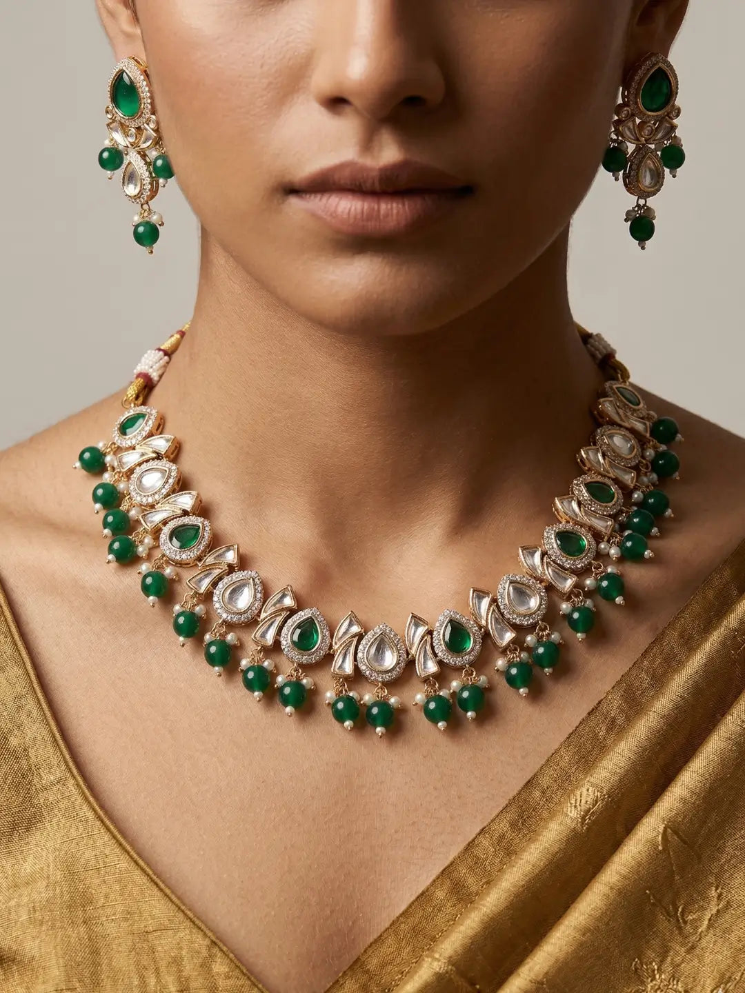 A close-up of a model wearing a gold-toned traditional necklace and matching drop earrings with emerald green stones and clear polki accents, paired with a gold saree.