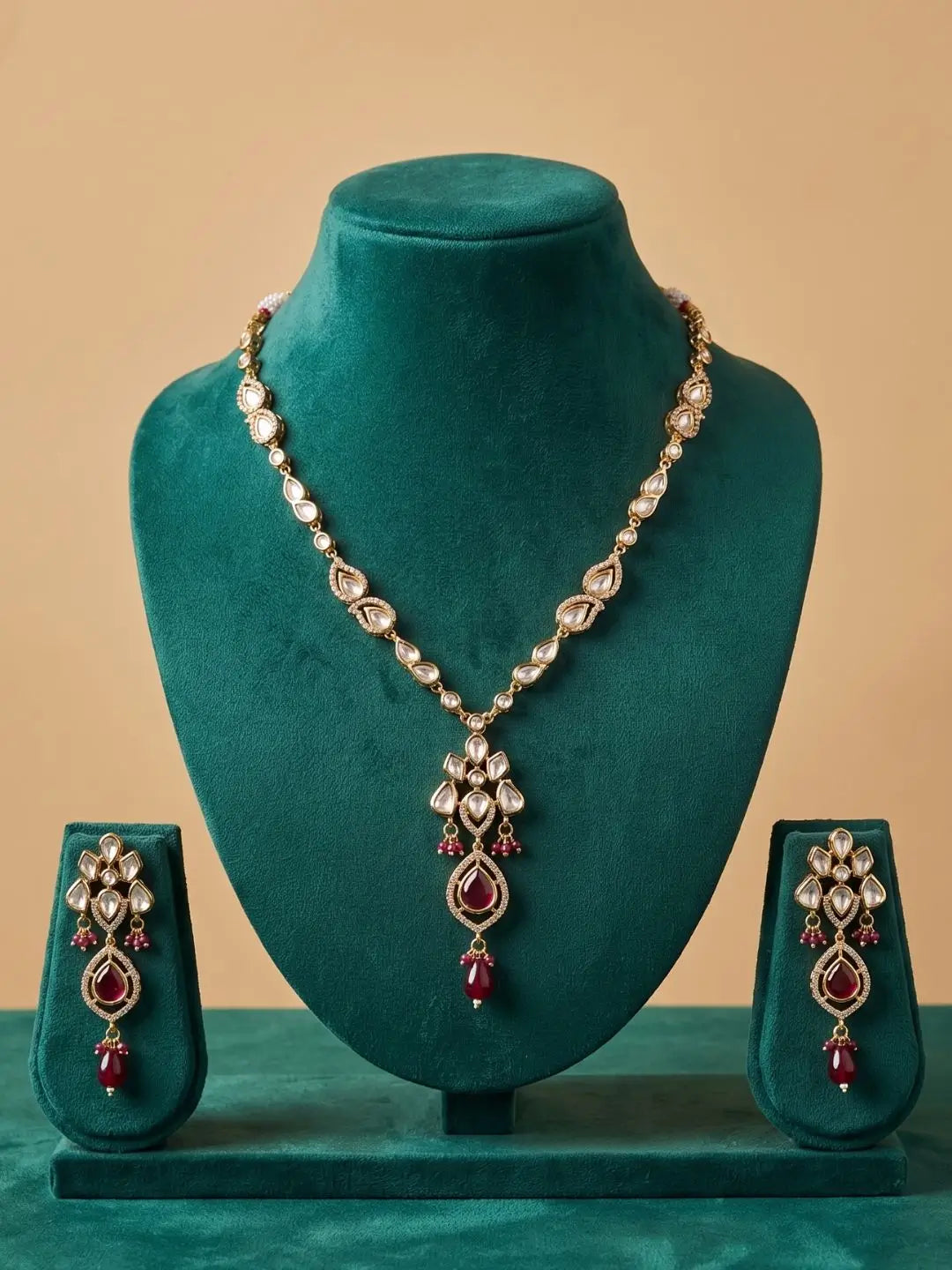 Gold-toned jewelry set with a necklace and matching earrings displayed on a dark teal velvet jewelry stand against a warm beige background. The jewelry features clear stones and red teardrop accents.