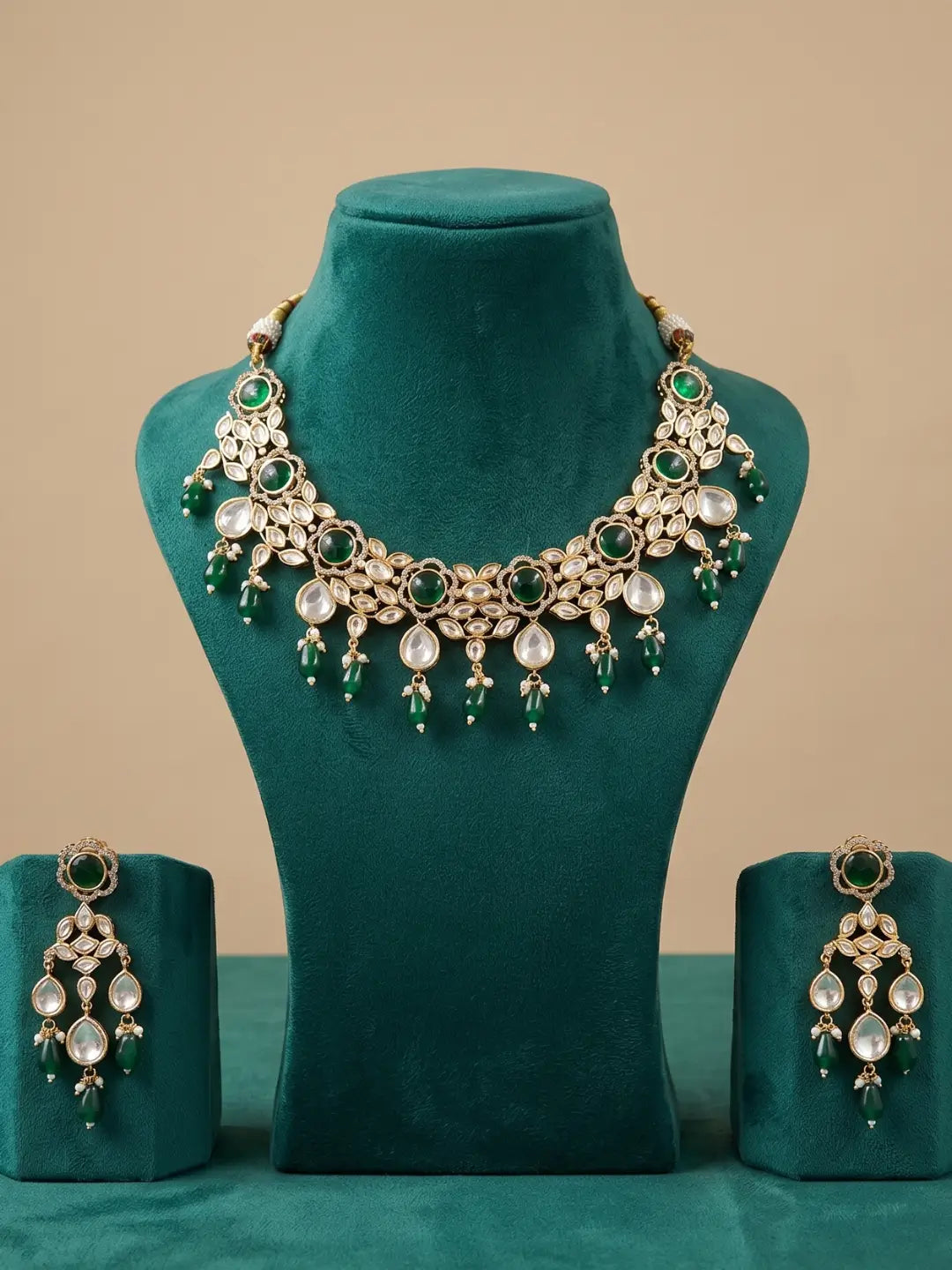 An ornate gold-toned kundan jewelry set displayed on a teal velvet mannequin bust, including a statement necklace and matching drop earrings with green beade drops.