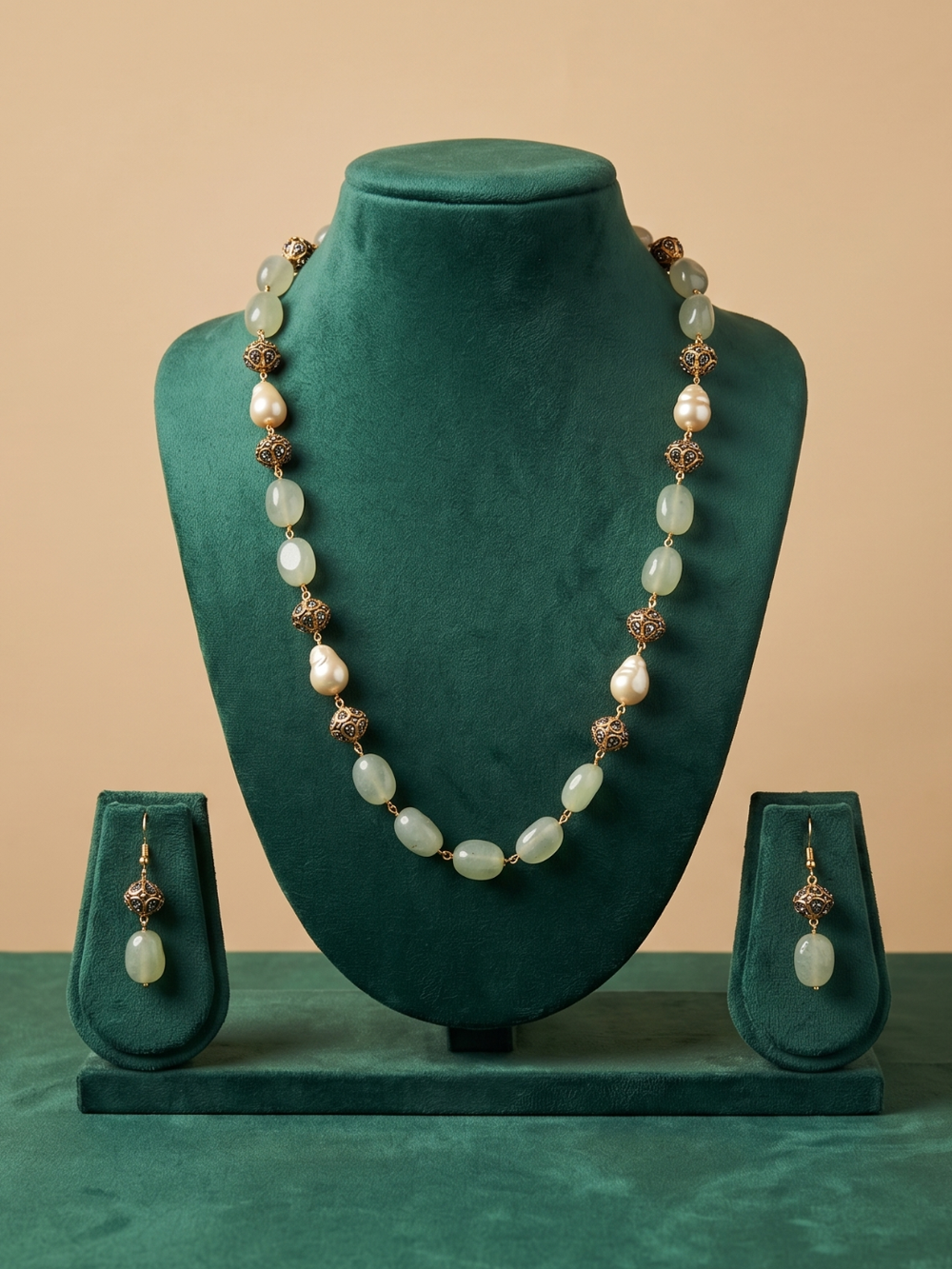 A jewelry set displayed on a dark green velvet mannequin bust, featuring a long beaded necklace and matching drop earrings. The beads are a mix of pale greenish-white pearls and metallic bronze-toned spacer beads, set against a solid beige background.
