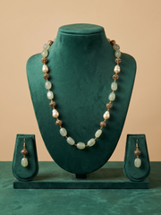 A jewelry set displayed on a dark green velvet mannequin bust, featuring a long beaded necklace and matching drop earrings. The beads are a mix of pale greenish-white pearls and metallic bronze-toned spacer beads, set against a solid beige background.