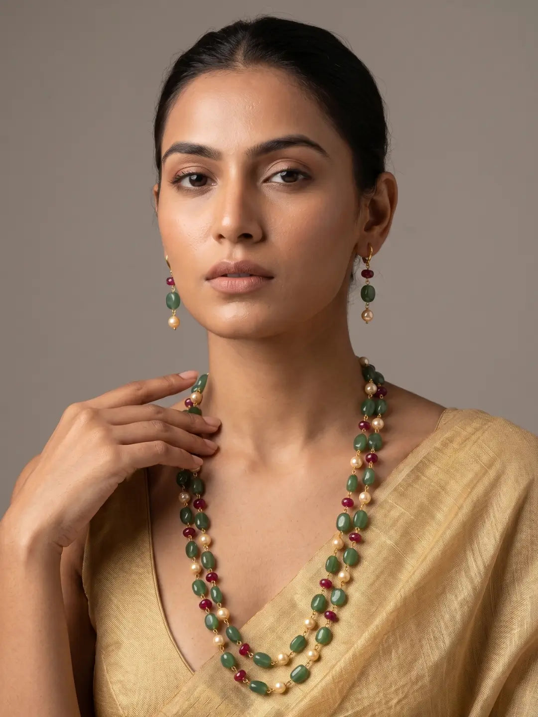 A woman wearing a double-strand beaded necklace and matching drop earrings. The jewelry features green oval beads, cream pearl-like beads, and small red beads, styled with a gold blouse against a neutral gray background.