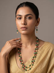 A woman wearing a double-strand beaded necklace and matching drop earrings. The jewelry features green oval beads, cream pearl-like beads, and small red beads, styled with a gold blouse against a neutral gray background.