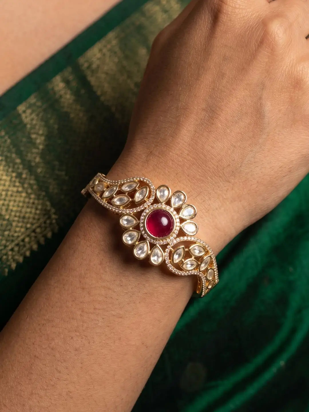 A gold-plated floral design kundan polki kada bracelet shown worn on a person's wrist against a green fabric background.