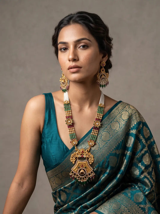 A woman wearing an ornate traditional Indian antique gold jewelry set with a long gold necklace, matching earrings, and a teal blue saree with gold zari work, posed against a neutral gray studio background.
