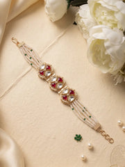 A traditional Indian kundan polki pearls bracelet featuring white faux pearls, gold-plated settings, red stones, and small green stones, displayed on a beige surface with white flowers and satin.