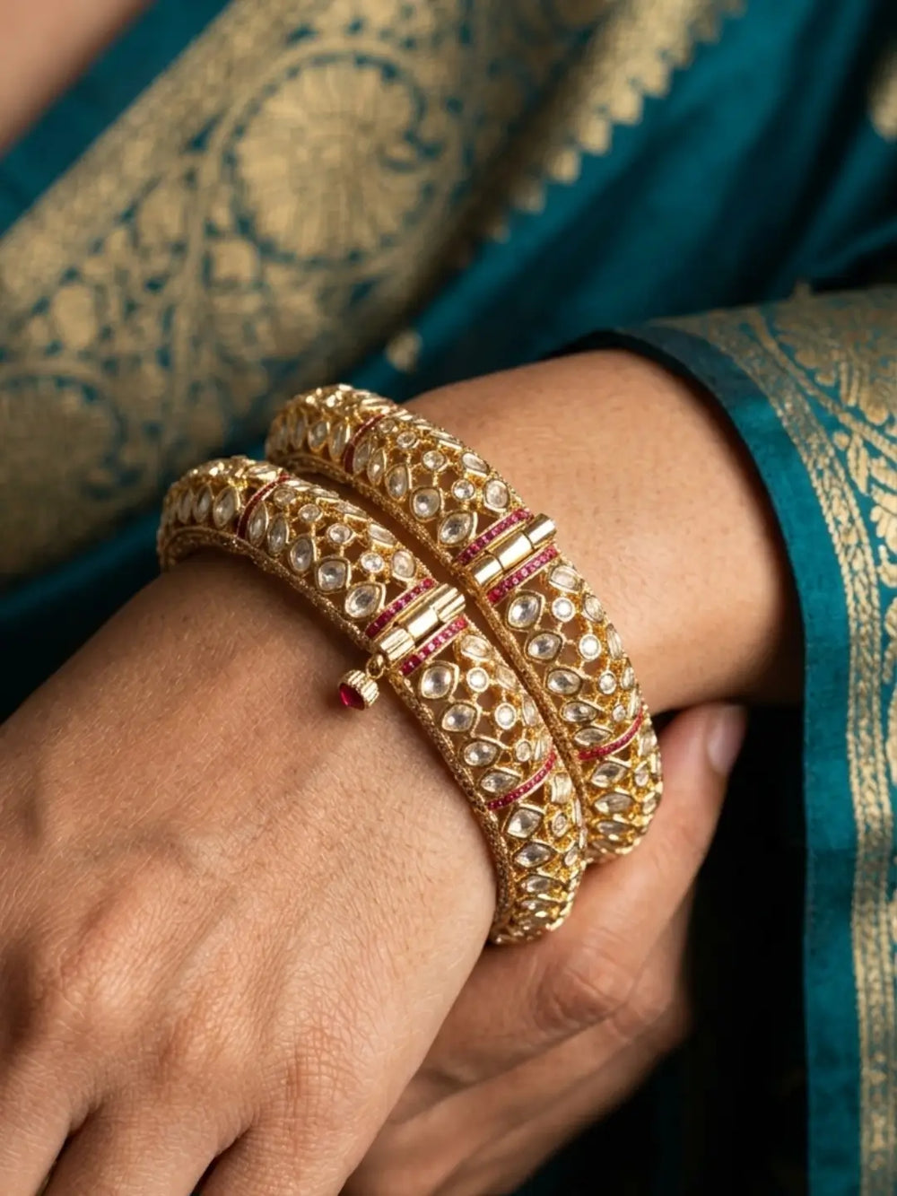 A close-up of a person's wrist wearing two stacked gold-toned red uncut polki bangles adorned with clear stones and red accents, set against a teal and gold patterned fabric background.