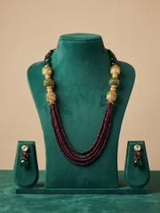 A traditional Indian beaded jewelry set displayed on a green velvet stand, featuring a multi-strand necklace with deep red beads, gold-toned ornate beads, and green accents, along with matching drop earrings.
