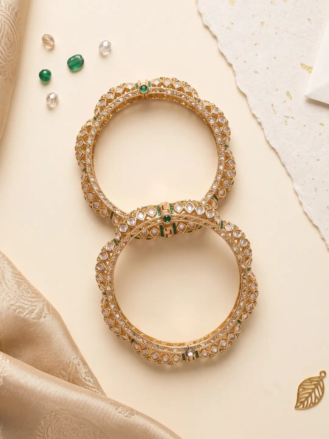 Two ornate gold-toned bangles decorated with clear stones and small green accents, arranged on a warm cream surface with decorative fabric and pearl-like beads.