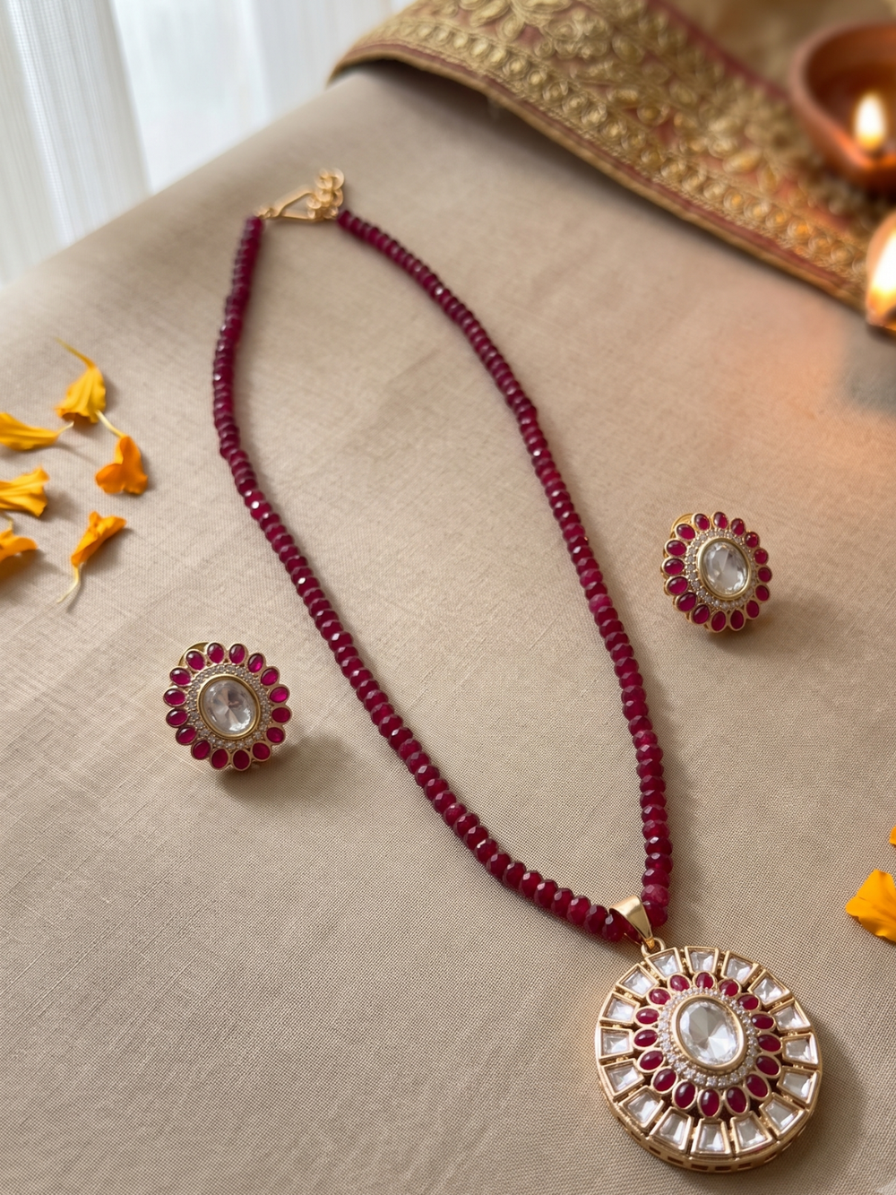 A traditional Indian jewelry set featuring a beaded necklace, a matching pendant, and earrings. The necklace and earrings are decorated with deep red beads and gold-plated metal borders, with a clear central stone.
