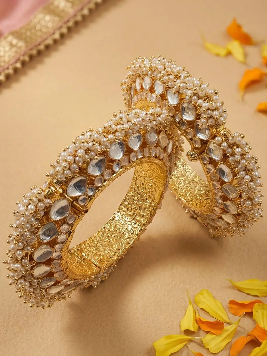 Two ornate gold-plated paachi kundan bangles decorated with white faux pearls and clear stones, arranged on a beige surface with scattered yellow and orange petals.