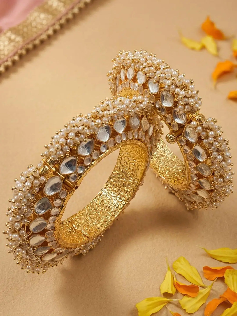 Two ornate gold-plated paachi kundan bangles decorated with white faux pearls and clear stones, arranged on a beige surface with scattered yellow and orange petals.