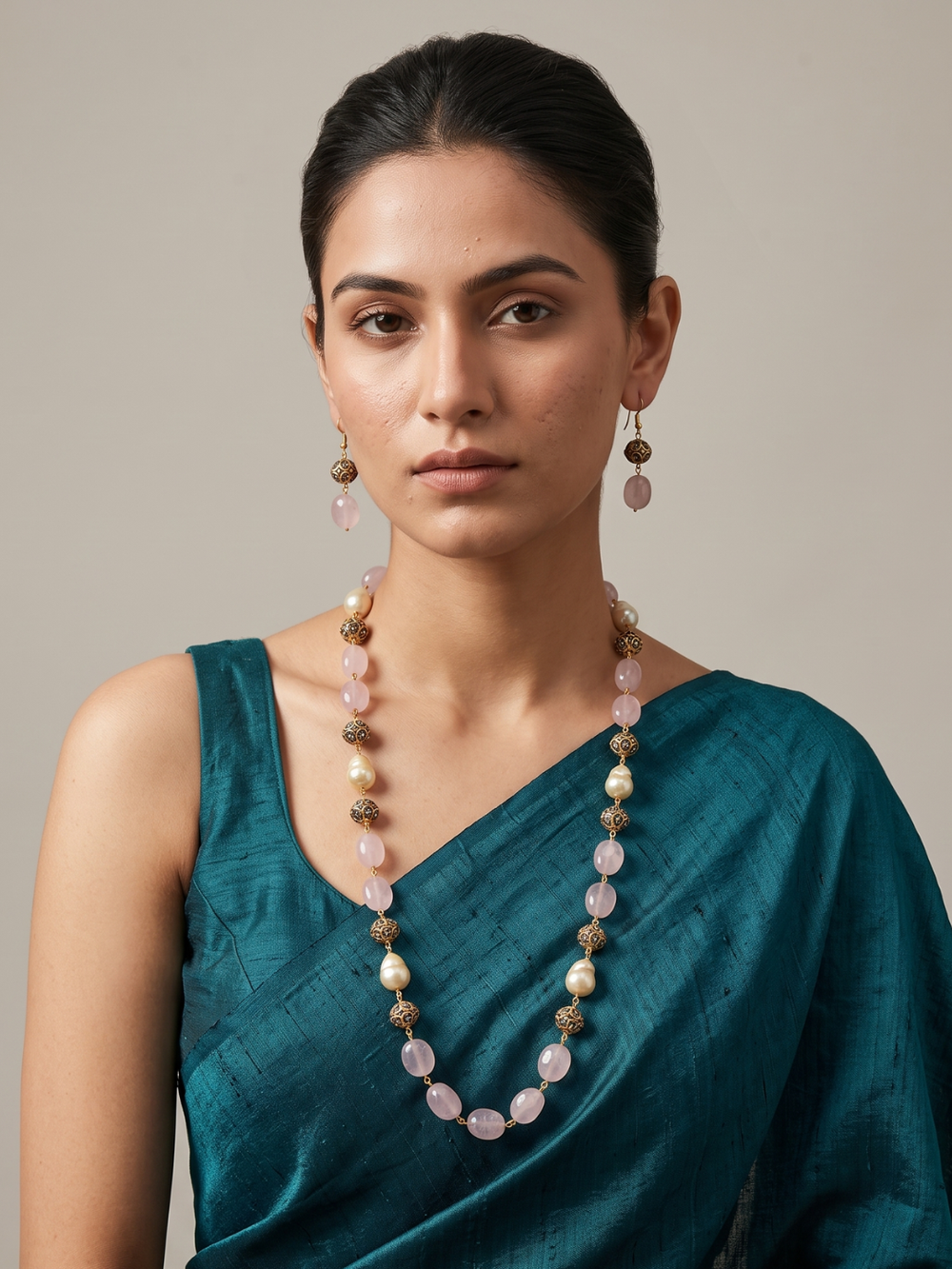 A woman wearing a long pink stone-beaded necklace and matching drop earrings, styled with a teal sari against a neutral beige background.