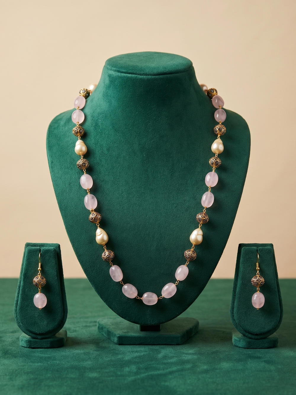 A beaded jewelry set displayed on a dark green velvet mannequin bust and matching stands, featuring a long necklace with pale pink oval beads, white pearl-like beads, and bronze-toned spacer details, plus matching drop earrings.