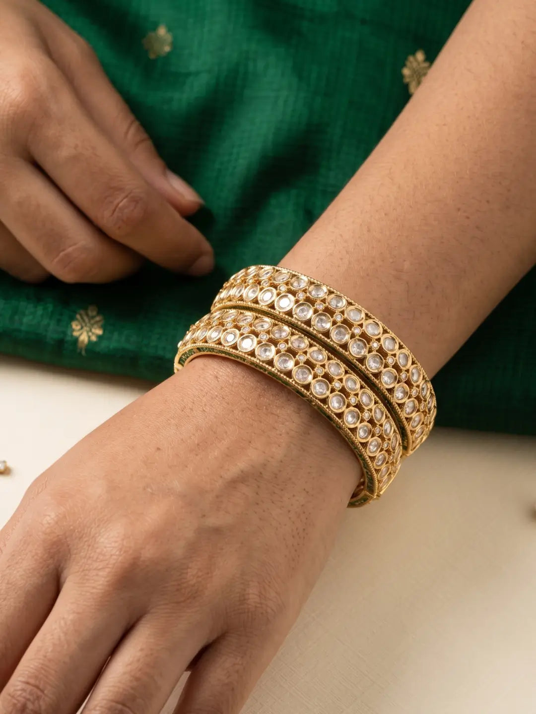 A close-up of a person's wrist wearing stacked gold plate polki bangles adorned with clear stones, set against a green embroidered garment and a light surface.