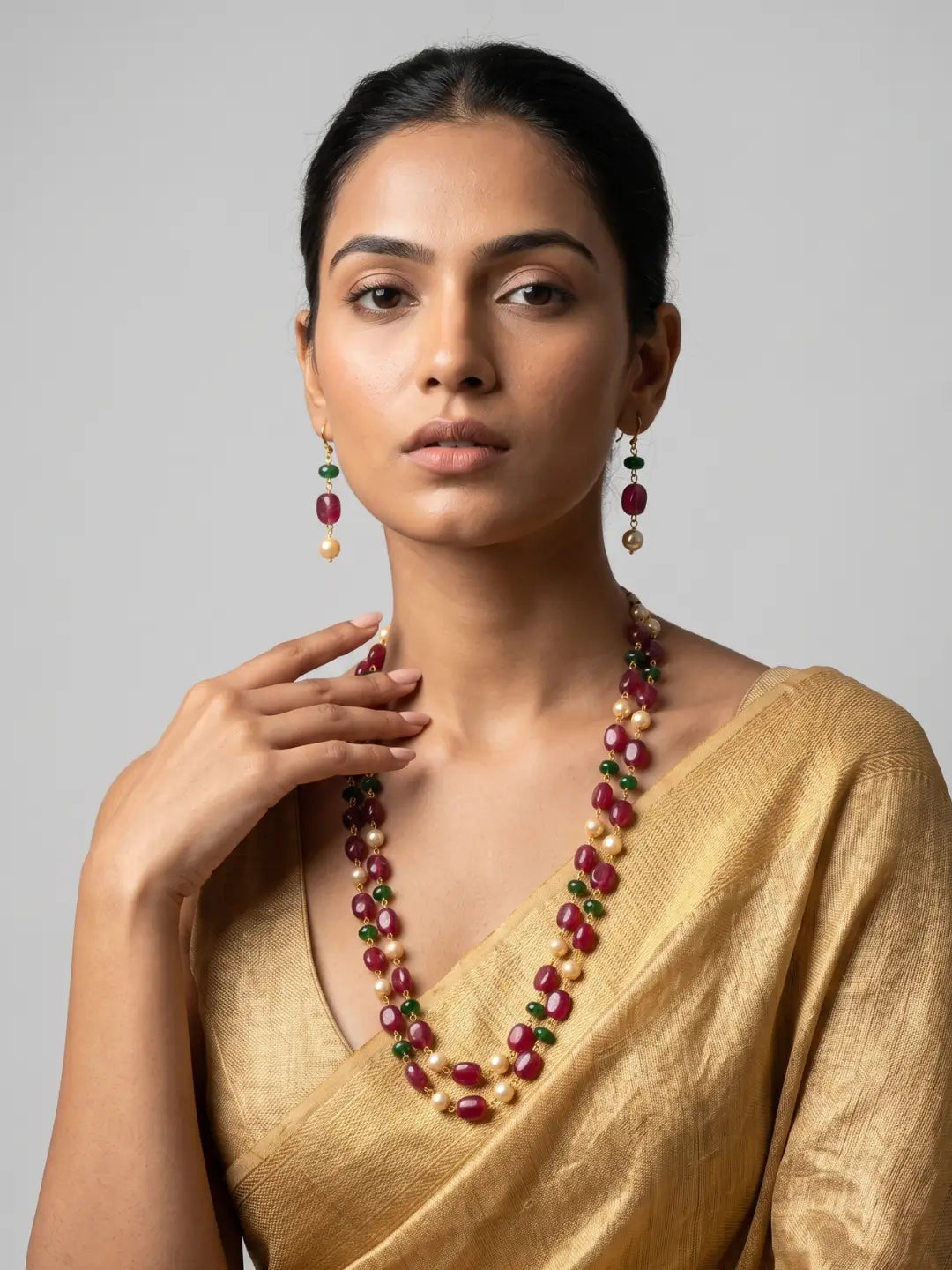A woman wearing a gold saree and matching statement jewelry, featuring a multi-strand necklace and matching drop earrings with deep red, green, and cream beads, set against a plain light gray background.