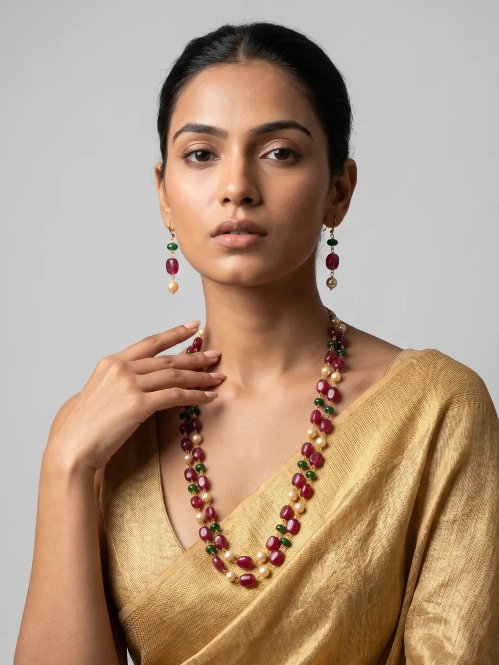 A woman wearing a gold saree and matching statement jewelry, featuring a multi-strand necklace and matching drop earrings with deep red, green, and cream beads, set against a plain light gray background.