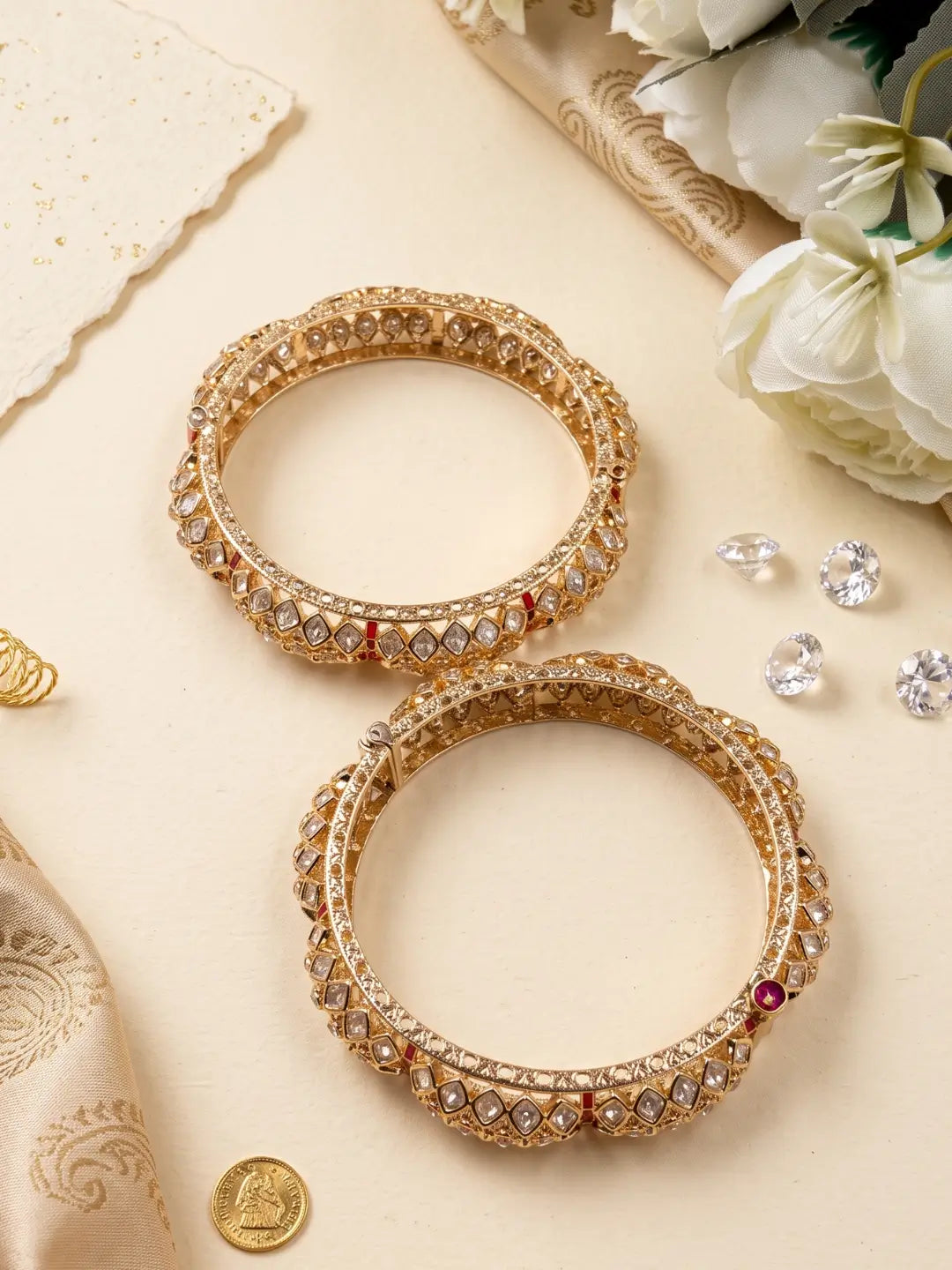 Two ornate gold-toned polki bangles with intricate crystal and red gemstone detailing, arranged on a beige surface with decorative fabric, flowers, and small clear stones.