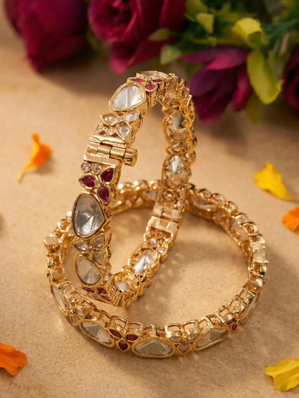 Two ornate gold-toned bangles studded with clear polki kundan stones and small red gem accents, arranged on a beige surface with blurred flowers in the background.