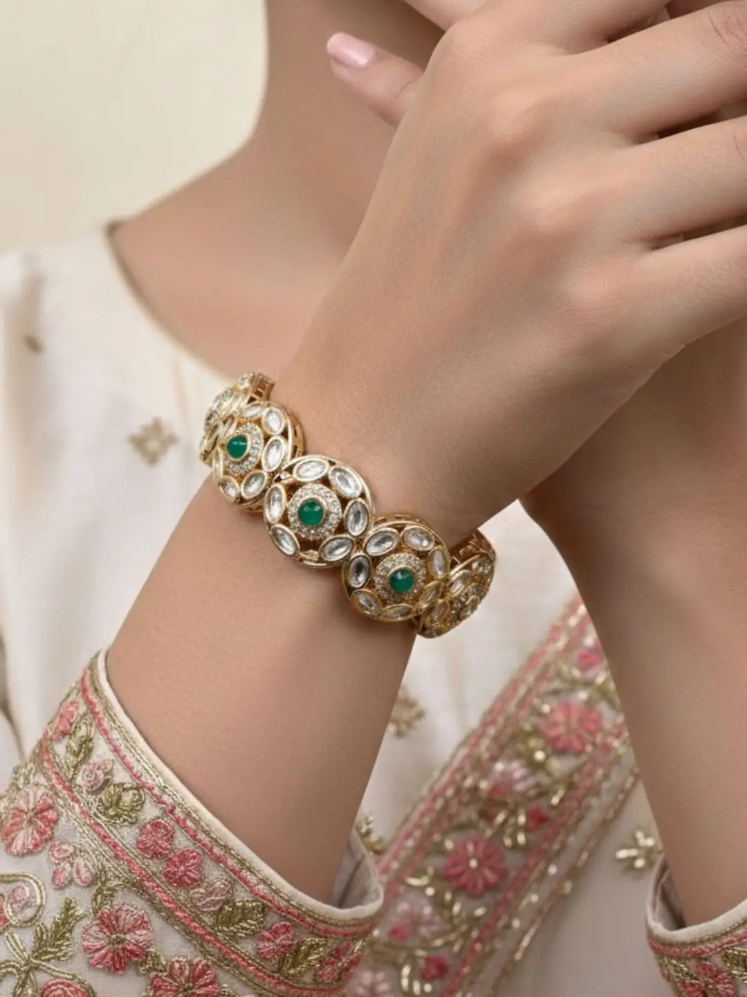 A close-up of a person's wrist wearing an ornate gold-toned traditional bracelet with green stone accents and clear stones, paired with a cream embroidered sleeve.