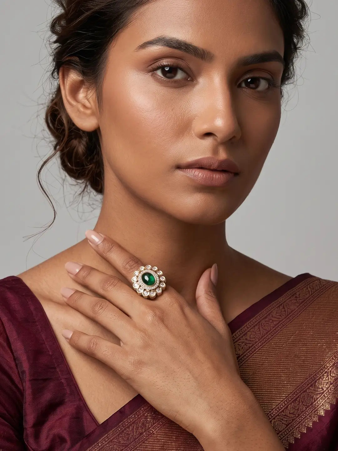 A close-up of a woman wearing a large statement ring with a green center stone and a halo of small clear polki stones, styled with a deep maroon traditional outfit against a neutral gray background.