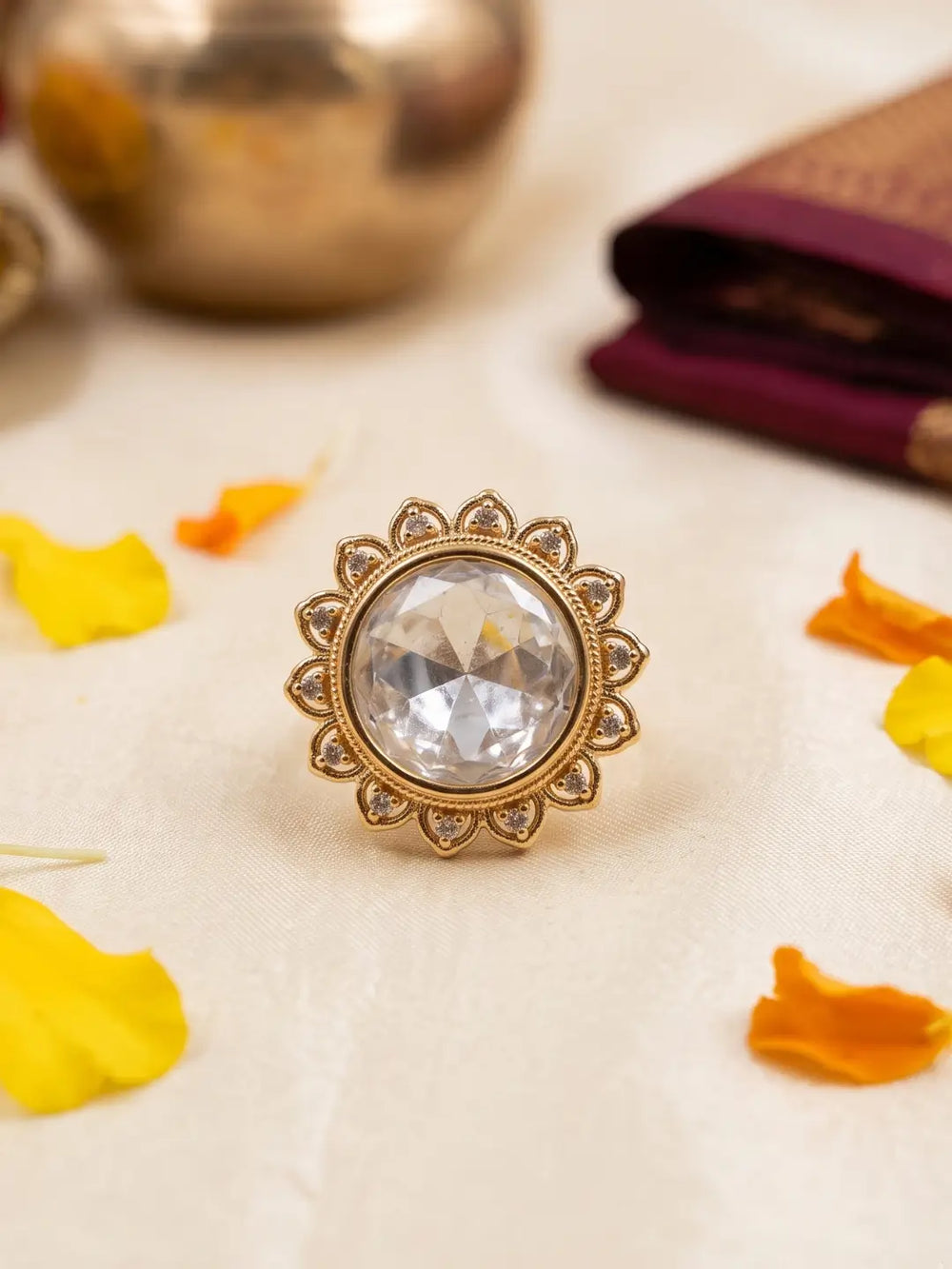A gold-toned kundan ring with a large clear faceted gemstone in a sunburst setting, styled on a cream fabric surface with scattered yellow and orange petals and blurred decorative objects in the background.