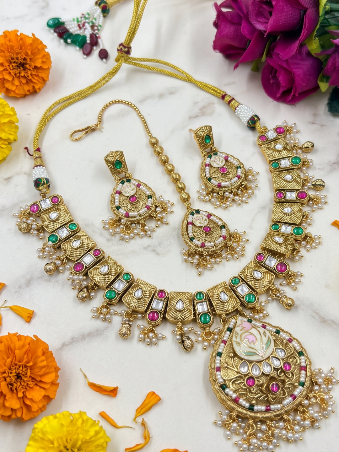 An ornate gold-plated Indian antique kundan jewelry set including a necklace, earrings, and a bracelet, decorated with faux pearls, green and pink stones, and red accents, arranged on a white marble surface with orange and purple flowers.