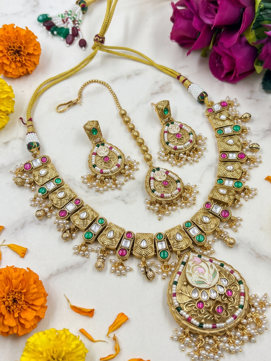 An ornate gold-plated Indian antique kundan jewelry set including a necklace, earrings, and a bracelet, decorated with faux pearls, green and pink stones, and red accents, arranged on a white marble surface with orange and purple flowers.