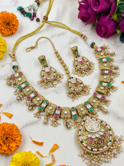 An ornate gold-plated Indian antique kundan jewelry set including a necklace, earrings, and a bracelet, decorated with faux pearls, green and pink stones, and red accents, arranged on a white marble surface with orange and purple flowers.