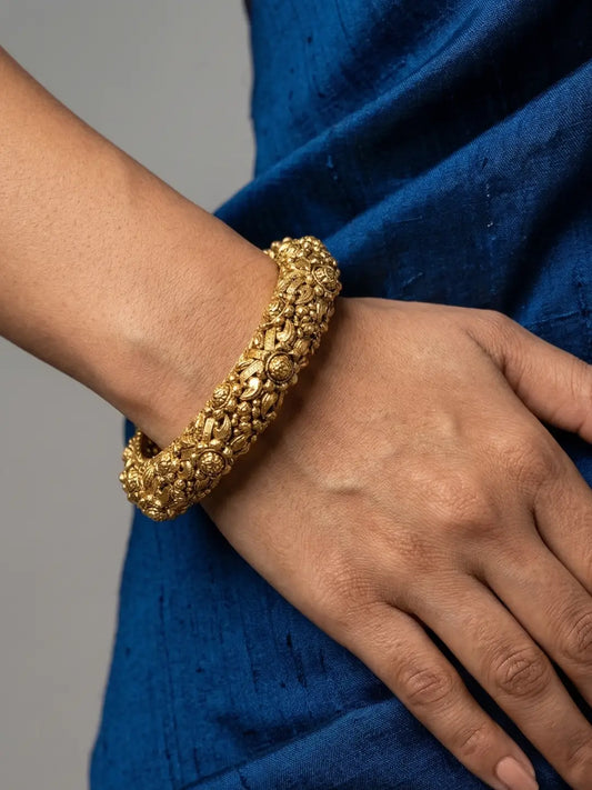 A close-up of a person's wrist and hand wearing a gold-plated antique golden nakshi kada bangle set against a blue fabric garment and a neutral gray background.