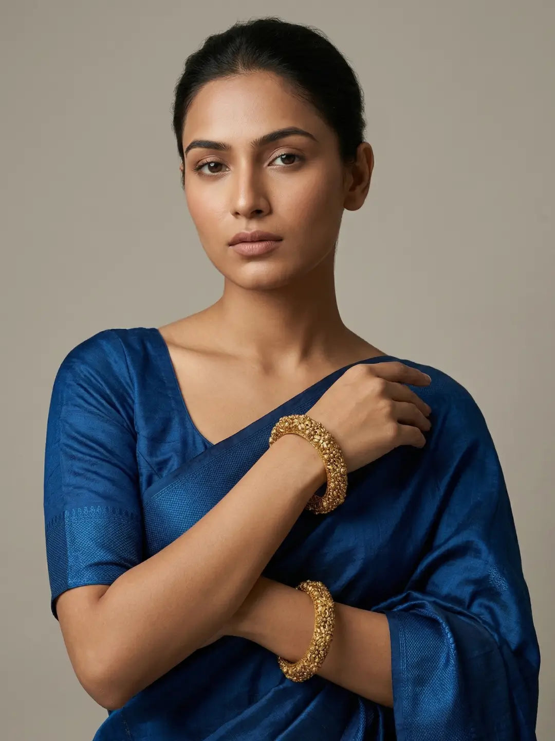 A woman wearing a royal blue satin saree and matching blouse, accessorized with two textured gold-plated antique bangles, posed against a neutral beige background.