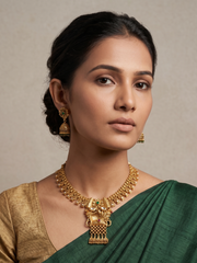 A woman wearing a gold-toned traditional antique gold necklace with a large pendant and matching earrings, paired with a green and gold sari, against a neutral beige background.