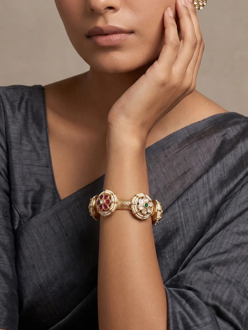 A close-up of a woman wearing a gold-toned multi-piece bracelet with red and green stone accents, paired with a dark gray draped blouse against a neutral beige background.