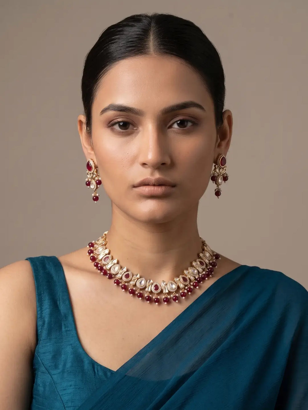 A woman wearing a gold-toned statement kundan necklace and matching drop earrings with deep red gemstone accents, paired with a teal blue blouse, set against a solid warm beige background.