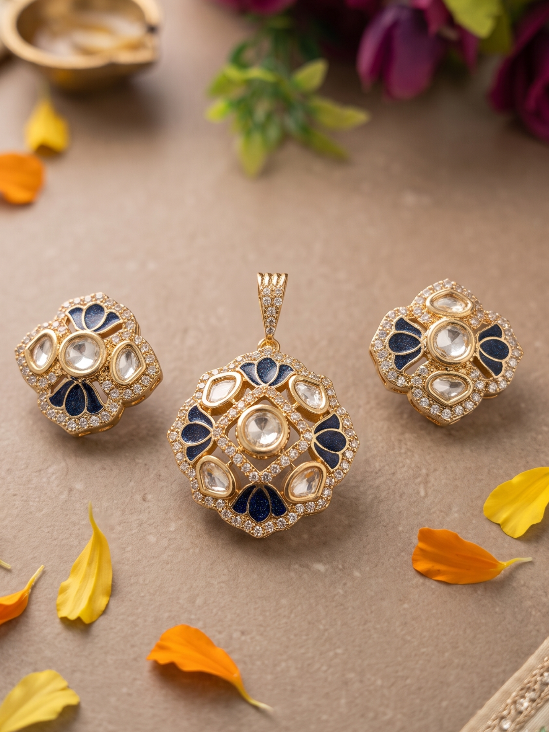 Gold-tone Indian Kundan jewelry set featuring a matching pendant and two matching earrings, decorated with clear stones, white crystals, and navy blue enamel details, displayed on a textured beige surface with scattered orange and yellow flower petals.