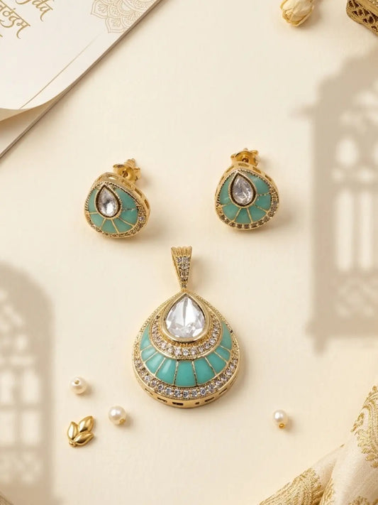 Gold-plated jewelry set featuring a teardrop-shaped pendant and matching earrings with turquoise green enamel, clear teardrop gemstones, and small crystal accents, arranged on a cream background with decorative shadows and gold-toned props.