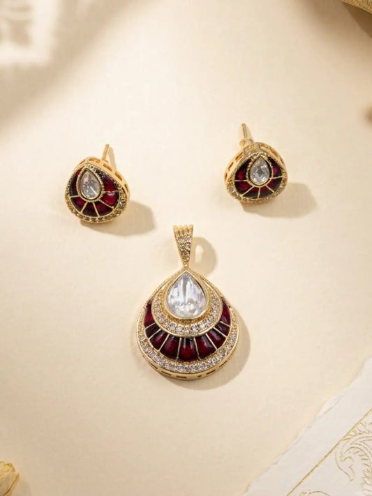 Gold-toned kundan jewelry set featuring a teardrop pendant and matching stud earrings with a central clear teardrop stone, surrounded by red enamel and white stone accents, displayed on a light beige surface.