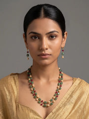 A woman with dark hair pulled back wears a double-strand beaded necklace and matching drop earrings. The jewelry features green stone beads, cream pearl-like beads, and small red accents, paired with gold-toned metal.