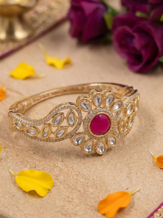 Decorative gold-plated bracelet with a red gemstone and polki stones on a beige surface with flowers in the background