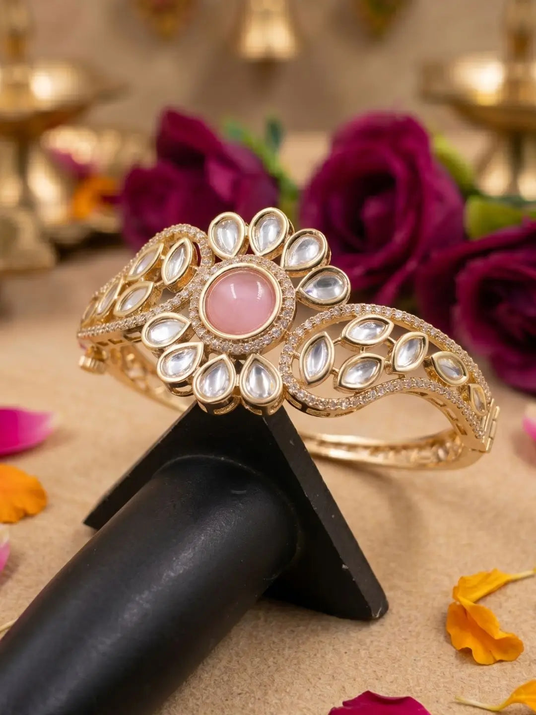 Gold-toned mandarin bangle bracelet with a central pink stone and clear stones in a floral pattern, displayed on a black stand with roses and gold diya lights in the background.