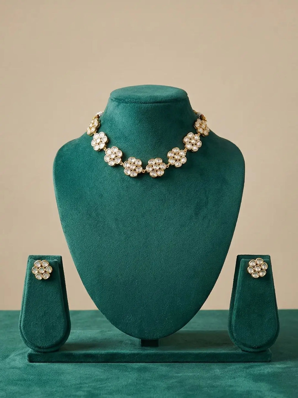 A floral statement polkinecklace and matching stud earrings displayed on a teal velvet jewelry bust set against a warm beige background.