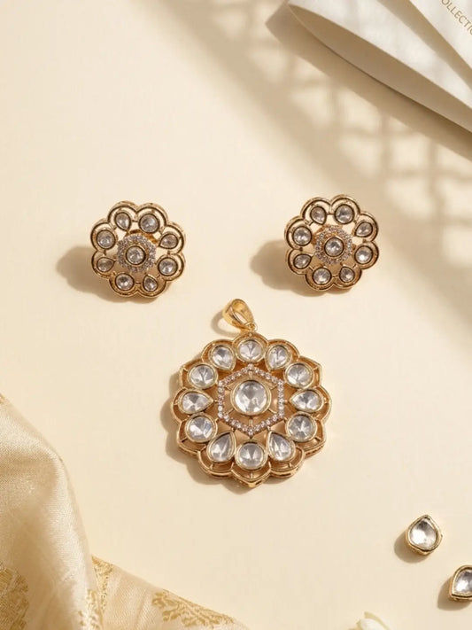 Gold-toned traditional kundan jewelry set featuring a pendant, matching small stud earrings with clear polki stones, arranged on a warm cream surface with soft shadows and decorative fabric.