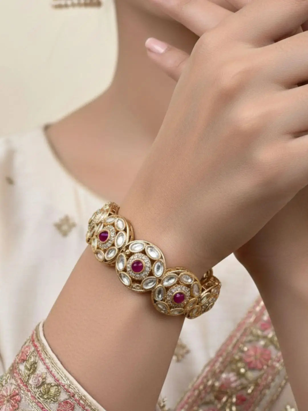 A close-up of a person wearing a gold-toned traditional Indian kundan  bracelet (bangle) embellished with white stones and red gemstones, paired with a cream-colored embroidered outfit.