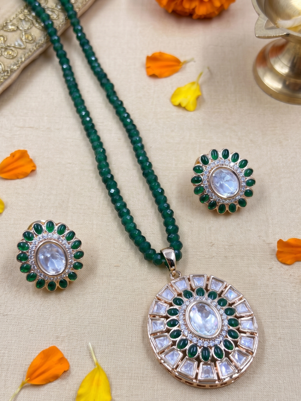 A green beaded Kundan jewelry set featuring a round pendant necklace, a matching stud earring,  arranged on a beige fabric background with orange flower petals.