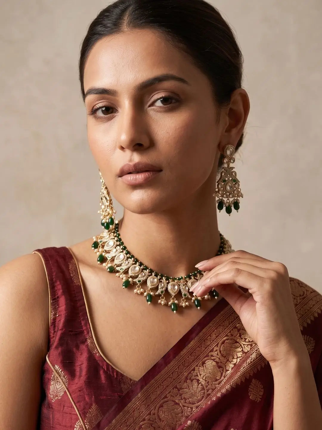 A woman wearing an ornate gold-toned polki necklace and matching drop earrings with green stone accents, paired with a deep maroon sari against a neutral beige background.